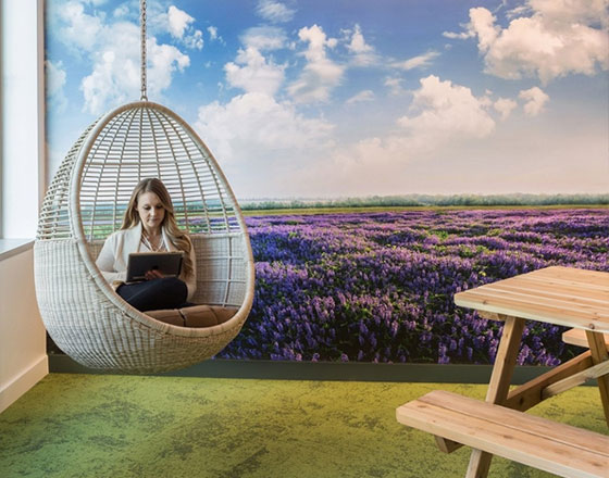 room with a mural of a lilac field with green carpet and a swing in the corner with a woman using a laptop