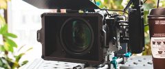 A professional film camera with a microphone attachment sits next to a coffee cup on an outdoor table surrounded by foliage