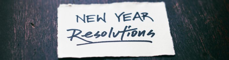 A handwritten note reading “New Year Resolutions” on a small piece of white paper placed on a dark wooden surface